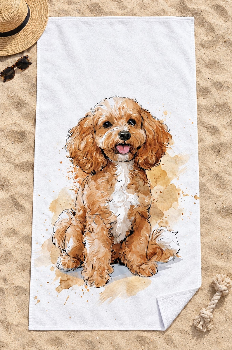 Top-down view of the whole watercolor Cavapoo beach towel laid flat on sand, the seated puppy surrounded by plain white space, with a straw hat, sunglasses and a rope toy; one corner folds back to the white back.