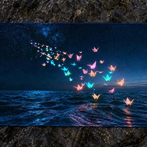 May include: A rectangular towel featuring a dark night sky filled with stars above a deep blue ocean. A swarm of glowing, multi-coloured origami paper cranes in shades of pink, orange, and blue floats across the scene, with some resting on the water.