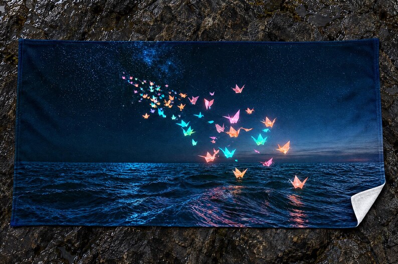 May include: A rectangular towel featuring a dark night sky filled with stars above a deep blue ocean. A swarm of glowing, multi-coloured origami paper cranes in shades of pink, orange, and blue floats across the scene, with some resting on the water.