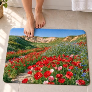 Red poppy field bath mat on a tiled floor in front of a white bathtub, showing poppies and blue wildflowers along a sandy path below pale hills and blue sky; bare feet step onto the top edge nearest the tub.