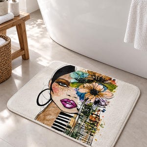 May include: A rectangular bath mat featuring a stylized illustration of a face with dark hair, bright pink lips, and a large hoop earring. The design incorporates watercolor-style floral elements in blue, orange, and green, with black stripes.