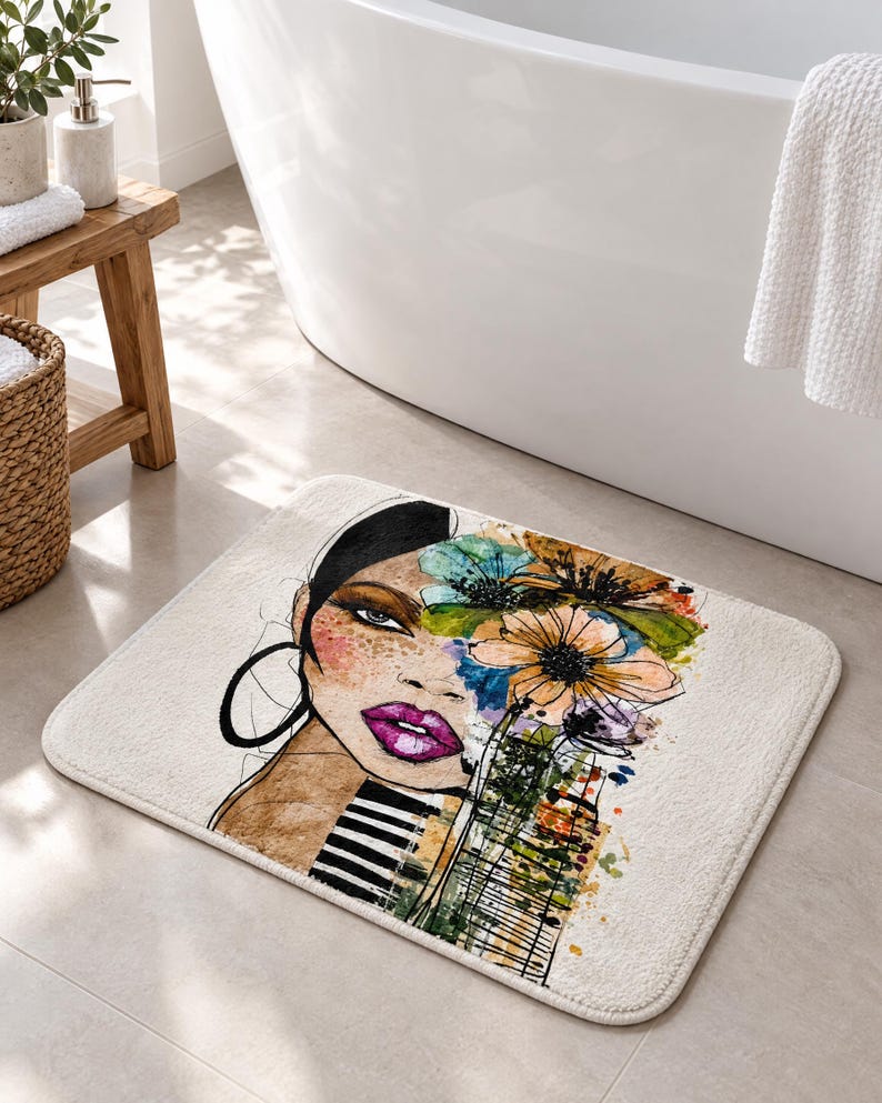 May include: A rectangular bath mat featuring a stylized illustration of a face with dark hair, bright pink lips, and a large hoop earring. The design incorporates watercolor-style floral elements in blue, orange, and green, with black stripes.