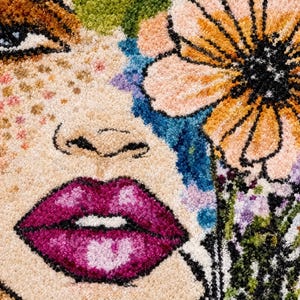 May include: A tufted rug featuring a close-up portrait of a face with freckles, vibrant magenta lips, and a large orange flower with black outlines. The design incorporates blue, green, and purple floral accents against a textured beige background.