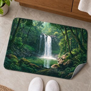 Jungle waterfall bath mat on a tiled floor by a wooden vanity and a potted plant; feet in white slippers rest on the tile in front and one corner flips up to show the gray anti-slip back.