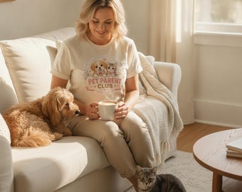May include: A cream-colored short-sleeve t-shirt featuring a graphic print of a puppy and a kitten inside a decorative frame with the text PET PARENT CLUB. The design includes small heart accents and floral elements surrounding the animals.