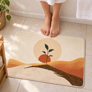 Boho sun sprout bath mat on a tiled floor in front of a white bathtub: a dark green sprout grows from a brown soil ridge before a terracotta half sun and pale circle, with ochre hills on cream; bare toes touch the top edge.