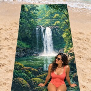 Jungle waterfall beach towel laid flat on sand by the surf, printed with a tall white waterfall falling into a green pool among mossy boulders and leafy trees; a woman in a coral swimsuit and sunglasses sits on the towel.