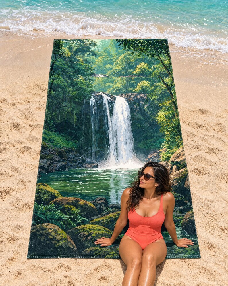 Jungle waterfall beach towel laid flat on sand by the surf, printed with a tall white waterfall falling into a green pool among mossy boulders and leafy trees; a woman in a coral swimsuit and sunglasses sits on the towel.