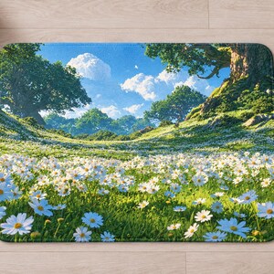 Overhead view of the whole white daisy meadow bath mat on a light wood floor, with rolled white towels, a lit candle and a small glass vase of daisies and purple wildflowers beside it.