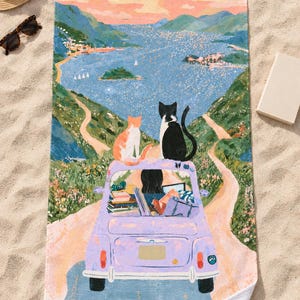 Top-down view of the whole two cats vintage car beach towel laid flat on sand, showing the dark-haired driver, the books and bags in the lavender car, the winding road and bay, with a straw hat and sunglasses.