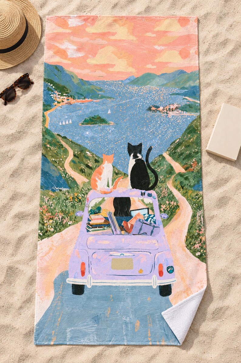 Top-down view of the whole two cats vintage car beach towel laid flat on sand, showing the dark-haired driver, the books and bags in the lavender car, the winding road and bay, with a straw hat and sunglasses.