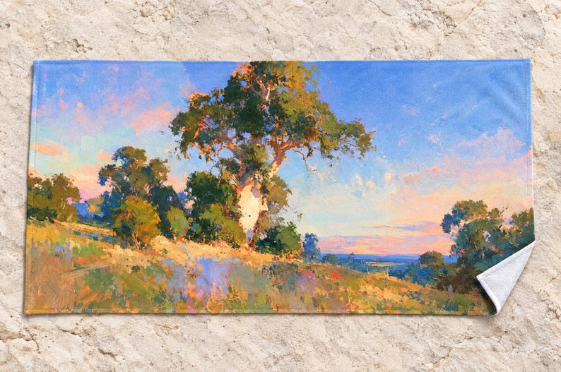 May include: A rectangular beach towel featuring a printed landscape painting of a large tree on a grassy hillside under a blue and pink sky. The artwork displays visible brushstrokes in shades of green, yellow, orange, and blue across the fabric.