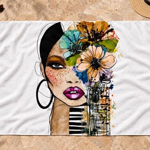 May include: A white rectangular beach towel featuring a stylized illustration of a face with dark hair, large hoop earrings, and bright pink lips. The design incorporates vibrant watercolor-style flowers in orange, blue, and green, alongside black and white stripes.