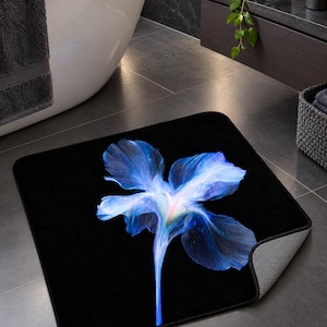 May include: A square bath mat with a solid black background featuring a large, vibrant blue iris flower graphic in the centre. The flower petals display shades of deep blue, light blue, and white, with a soft, glowing light effect at the base.
