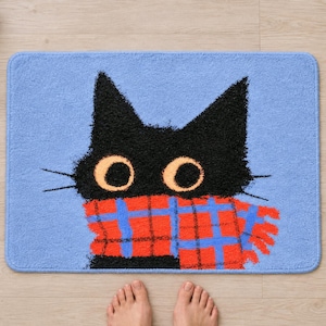 Plaid scarf black cat bath mat seen from above on a light wood floor, with rolled white towels, a candle, a pine sprig and bare feet at the bottom edge.