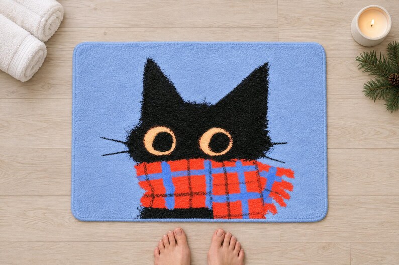 Plaid scarf black cat bath mat seen from above on a light wood floor, with rolled white towels, a candle, a pine sprig and bare feet at the bottom edge.