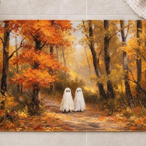 Puede incluir: Una alfombrilla de ba&ntilde;o rectangular con un estampado al estilo de pintura al &oacute;leo de dos fantasmas blancos de pie en un sendero forestal rodeado de &aacute;rboles oto&ntilde;ales de color naranja y amarillo con hojas ca&iacute;das en el suelo.