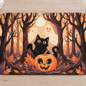 Overhead view of the whole black cat jack-o'-lantern bath mat on a light wood floor, with rolled white towels, a lit candle, a small pumpkin and a plant placed beside it.