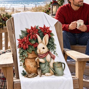 Poinsettia bunny beach towel draped over a wooden deck chair by the ocean, with a smiling man in a red sweater holding a mug beside it.