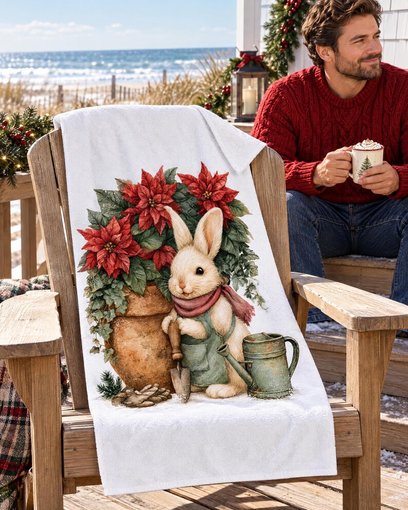 Poinsettia bunny beach towel draped over a wooden deck chair by the ocean, with a smiling man in a red sweater holding a mug beside it.