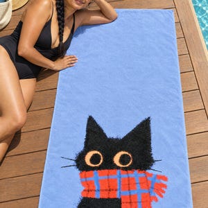 Plaid scarf black cat beach towel on a wooden pool deck, printed with a black cat's head and round eyes in a red plaid scarf on periwinkle blue, as a woman in a black swimsuit and sunglasses lies beside it.