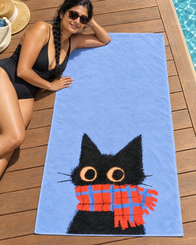 Plaid scarf black cat beach towel on a wooden pool deck, printed with a black cat's head and round eyes in a red plaid scarf on periwinkle blue, as a woman in a black swimsuit and sunglasses lies beside it.