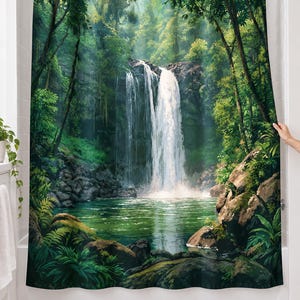Rainforest waterfall shower curtain over a bathtub in a white-tiled bathroom: a tall waterfall spills into a green pool between mossy boulders, ferns and jungle trees; a hand pulls back the right edge.