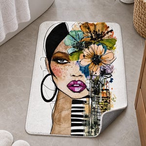 May include: A rectangular bath mat featuring a stylized illustration of a person's face with dark hair, bright purple lips, and large hoop earrings. The design includes colorful watercolor-style flowers and black and white striped patterns.
