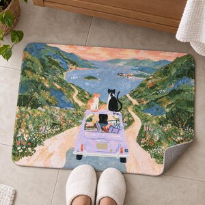 Cats road trip bath mat on a tiled floor by a wooden vanity, showing the lavender car, its dark-haired driver and two cats on the road to the bay; feet in white slippers rest in front and one corner flips up to show the gray back.