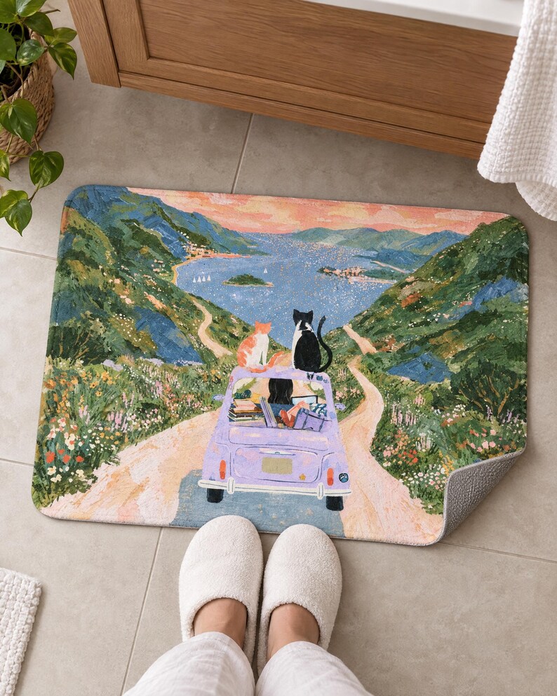 Cats road trip bath mat on a tiled floor by a wooden vanity, showing the lavender car, its dark-haired driver and two cats on the road to the bay; feet in white slippers rest in front and one corner flips up to show the gray back.
