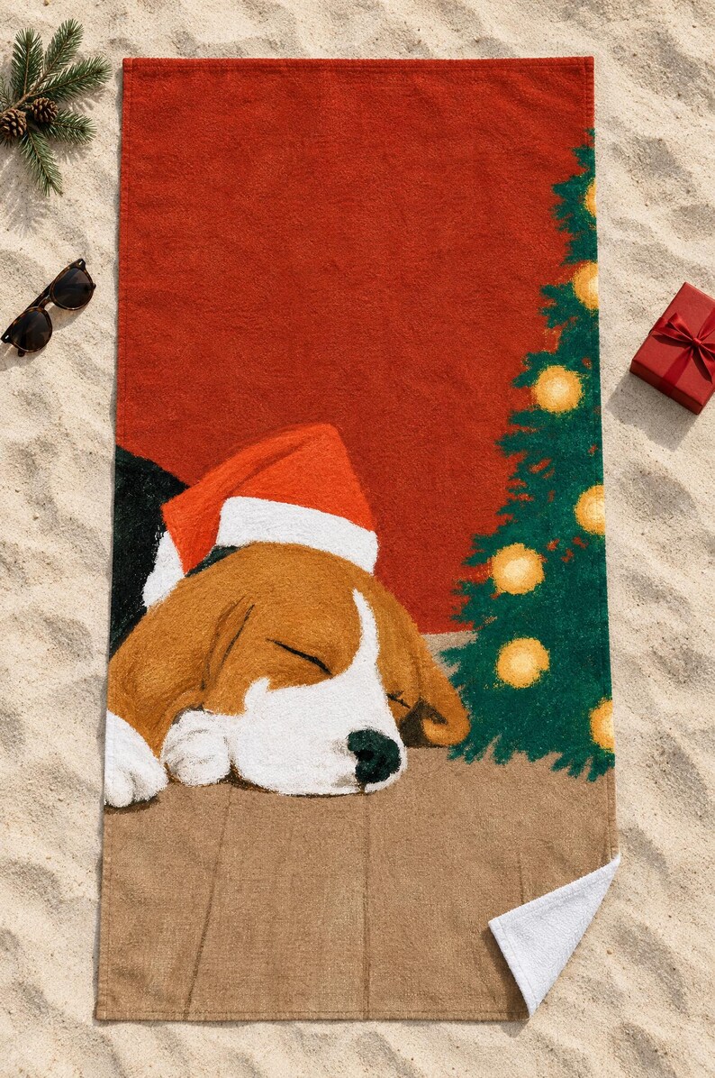 Sleeping beagle Santa hat beach towel laid flat on sand with sunglasses, a pine sprig and a small red gift box, one corner folded to show the white back.