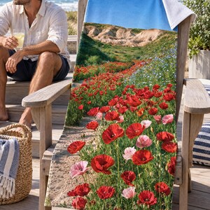 Red poppy field beach towel draped on a wooden Adirondack chair on a sunny beach deck, printed with red and pink poppies on a path toward hills and sky, as a man in sunglasses sits on the steps nearby.