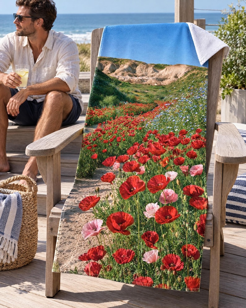 Red poppy field beach towel draped on a wooden Adirondack chair on a sunny beach deck, printed with red and pink poppies on a path toward hills and sky, as a man in sunglasses sits on the steps nearby.