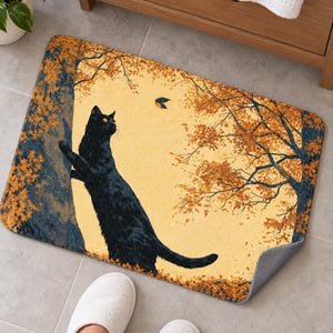 Bath mat with a cat stretching up an autumn tree toward a bird, on a gray tiled floor by a wooden vanity; feet in white slippers rest at the front and one corner flips up to show the gray anti-slip back.