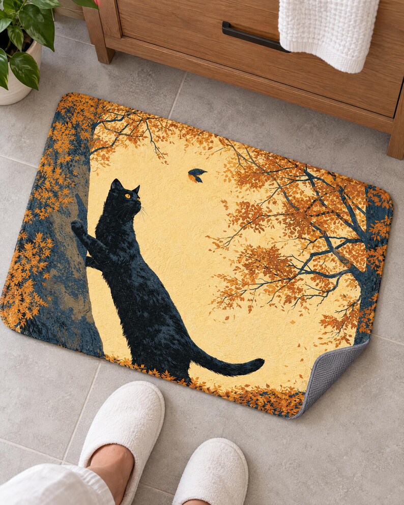 Bath mat with a cat stretching up an autumn tree toward a bird, on a gray tiled floor by a wooden vanity; feet in white slippers rest at the front and one corner flips up to show the gray anti-slip back.