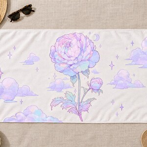 May include: A rectangular beach towel featuring a central large, pastel-colored peony flower with a smaller bud on a stem. The background is white with scattered light purple clouds, small four-pointed stars, and a crescent moon illustration.