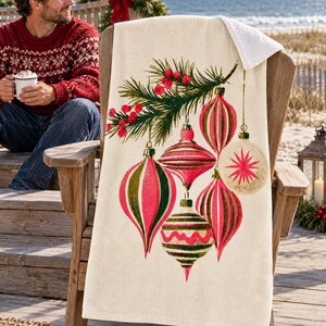 Vintage ornament beach towel draped over a wooden Adirondack chair on a seaside deck with a wreath, red bow and lantern; a top corner folds back to the white terry back, and a man in a red patterned sweater sits nearby with a mug of cocoa.