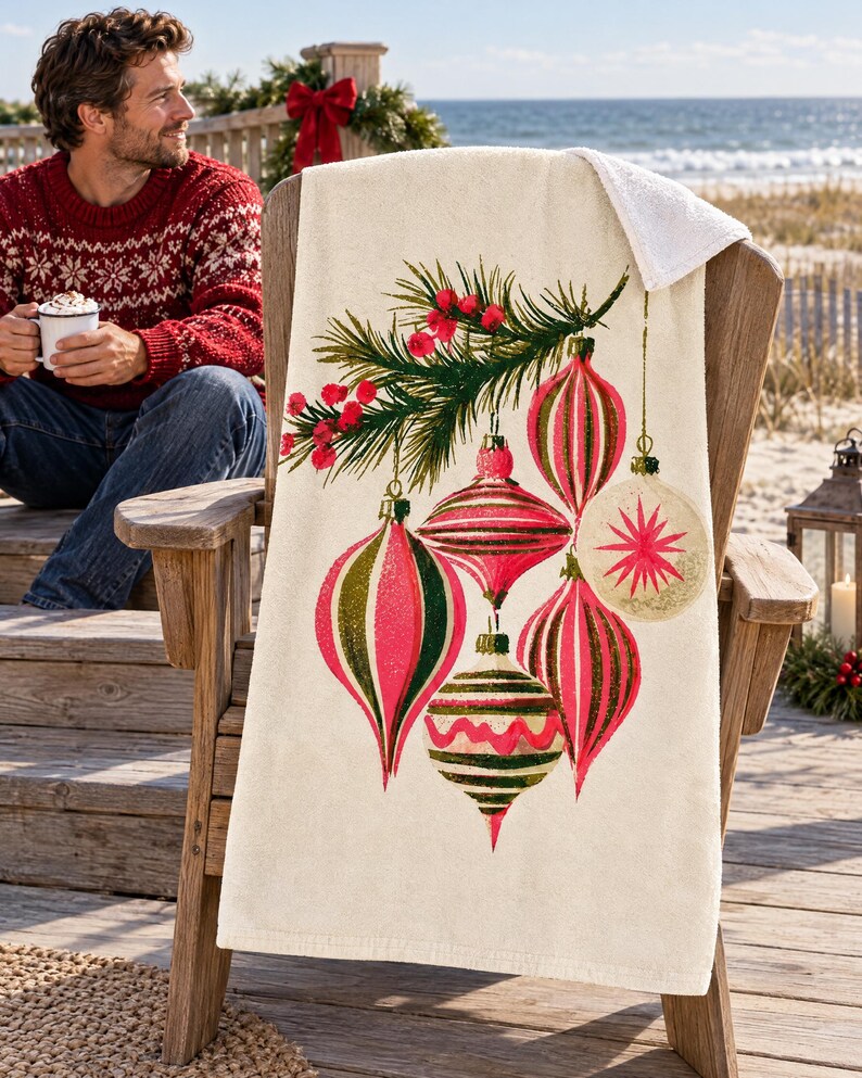 Vintage ornament beach towel draped over a wooden Adirondack chair on a seaside deck with a wreath, red bow and lantern; a top corner folds back to the white terry back, and a man in a red patterned sweater sits nearby with a mug of cocoa.