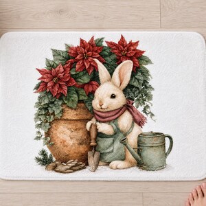 Poinsettia bunny bath mat seen from above on a light wood floor, with rolled white towels, a candle, a pine sprig and bare feet at the bottom edge.