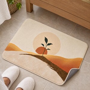 Neutral boho bath mat with a sprout, a terracotta sun and ochre hills on a tiled floor by a wooden vanity; feet in white slippers rest on the tile in front and one corner flips up to show the gray anti-slip back.