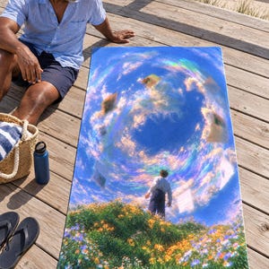 Surreal sky beach towel on a wooden boardwalk, printed with a figure on a flowery hill under a swirling rainbow-tinted sky with floating clouds, as a man in a blue shirt and shorts sits beside it.