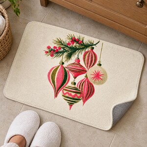 Cream bath mat with vintage glass ornaments and a berry pine sprig on a tiled floor by a wooden vanity and a basket; feet in white slippers rest at the front and one corner flips up to show the gray anti-slip back.