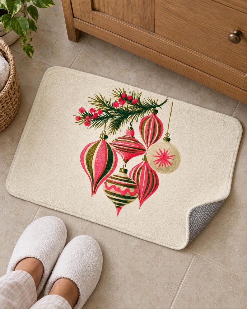 Cream bath mat with vintage glass ornaments and a berry pine sprig on a tiled floor by a wooden vanity and a basket; feet in white slippers rest at the front and one corner flips up to show the gray anti-slip back.