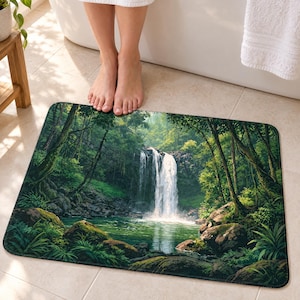 Jungle waterfall bath mat on a tiled floor in front of a white bathtub: a white waterfall pours into a green pool framed by tall trees, mossy rocks and ferns; bare feet stand on the top edge nearest the tub.
