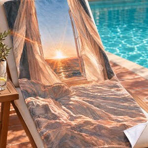 May include: A rectangular beach towel featuring a printed image of an open window looking out onto a sunset over the ocean. The design includes sheer white curtains blowing inward, a bright golden sun on the horizon, and rocky terrain in the foreground.