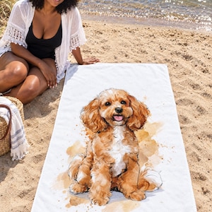 Cavapoo dog beach towel laid on sand by the water, printed with a fluffy apricot puppy sitting with its tongue out on white, as a smiling woman in a sun hat and white cover-up sits beside it.