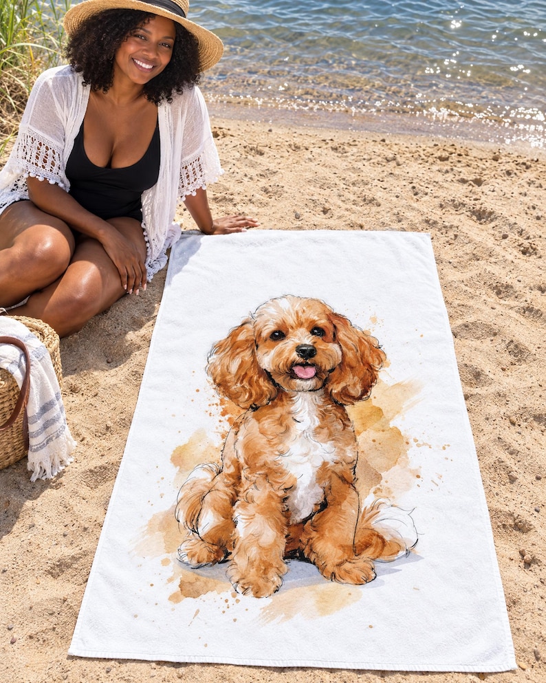 Cavapoo dog beach towel laid on sand by the water, printed with a fluffy apricot puppy sitting with its tongue out on white, as a smiling woman in a sun hat and white cover-up sits beside it.