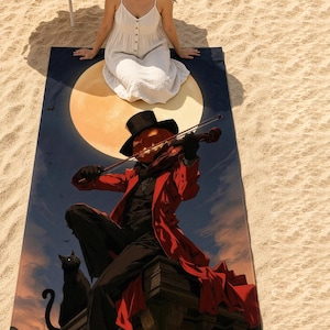 Halloween beach towel on sunny sand, with a woman in a white sundress sitting at the top edge. The print shows a pumpkin-head fiddler in a red coat playing violin on a rooftop under a full orange moon, with a black cat beside him.