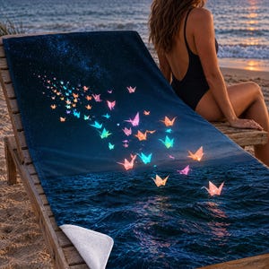 May include: A rectangular beach towel featuring a dark blue night sky transitioning into deep ocean waves. The design displays a swarm of glowing, multicoloured origami cranes in shades of pink, orange, and cyan, scattered across the surface.