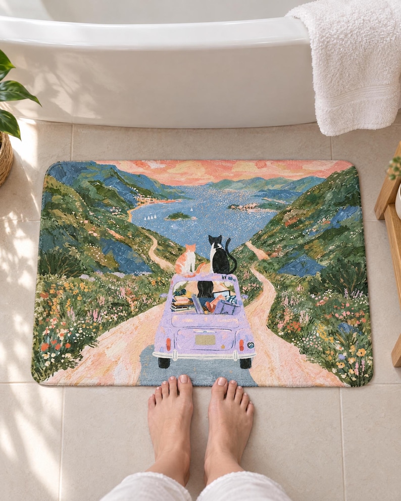 Cats road trip bath mat on a tiled floor in front of a white bathtub: two cats sit atop a lavender car as a dark-haired driver heads down a winding road toward a blue bay and green hills; bare feet stand on the tile below it.